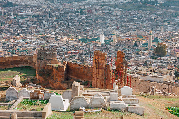 White gravestones scatter across a hillside cemetery near ancient fortress walls, with the city of Fez stretching beyond. The composition reflects spirituality, history, and continuity.