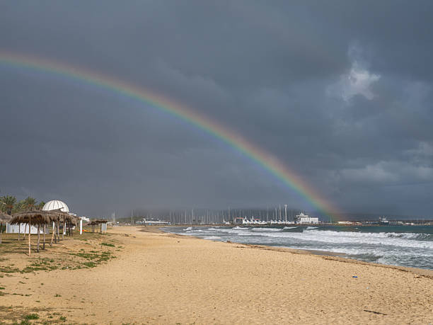 A stunning rainbow arches over the empty shores of Tunisia as a winter sun pierces through retreating rain clouds. The vibrant colors contrast with the deep blue Mediterranean Sea, offering a rare moment of serene beauty and solitude.
