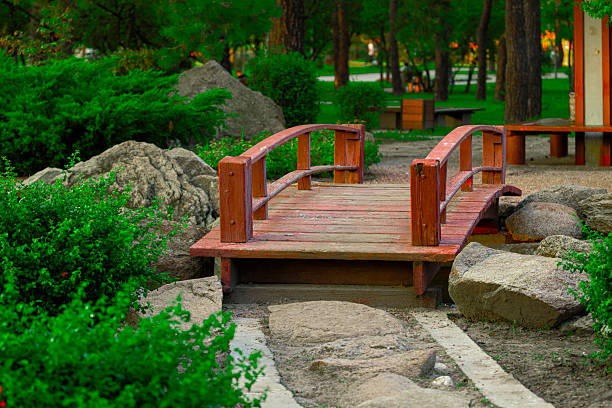 A charming red wooden bridge with curved railings invites passage across a dry creek bed in a peaceful park setting surrounded by vibrant foliage and natural stones