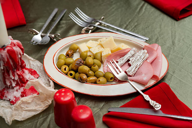 Close-up of an appetizer plate featuring green olives, cubes of cheese, cured salami sticks and folded ham, arranged on a rustic white plate with a red rim. The plate sits on an olive-green tablecloth with silver cutlery, red cloth napkins, and red-and-white dripping candles nearby, creating a cozy festive dining atmosphere.