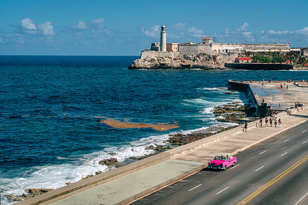Pink vintage convertible driving along the Havana Malecón beside crashing blue waves, with El Morro fortress and lighthouse on the rocky coastline and people strolling on the seaside promenade.