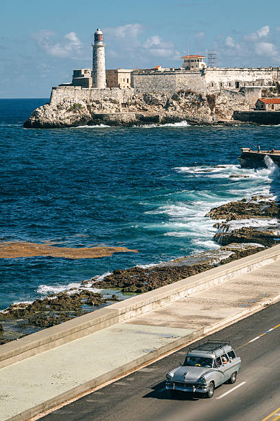 Classic American car drives along Havana's seaside Malecon with waves breaking on the rocky shore and a historic fortress and lighthouse in the background, conveying travel, nostalgia, and Cuban coastal life.