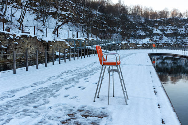 Orange lifeguard chair stands on a snow covered pier at zakrzowek quarry pool. Frozen surface, footprints, and quiet water create an off-season mood, emphasizing safety, closed swimming season, and winter recreation space.