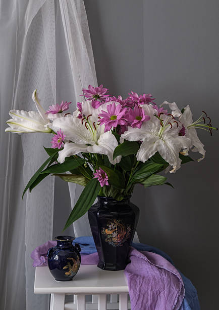 White Lilies And Pink Daisies Bouquet Soft Window Light Over Glass Vase On White Stool With Layered Pastel Drapery, Gentle Shadows, Glossy Green Leaves And Water, Intimate Studio