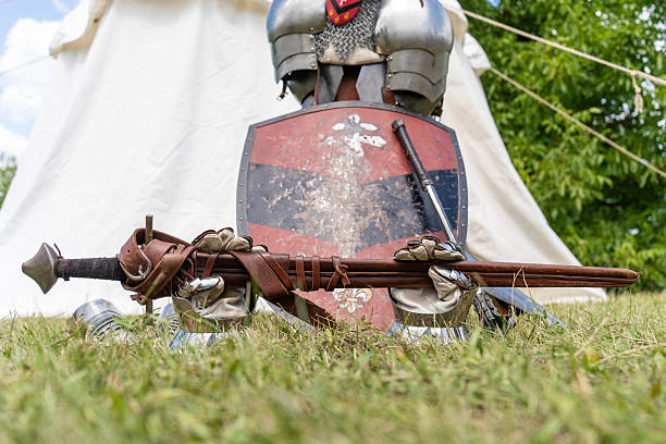 Close-up of a medieval sword and decorative shield resting on green grass, surrounded by steel gauntlets and armor pieces. The worn red and black shield with fleur de lis symbols and the leather-bound sword evoke historical reenactment and chivalric tradition.