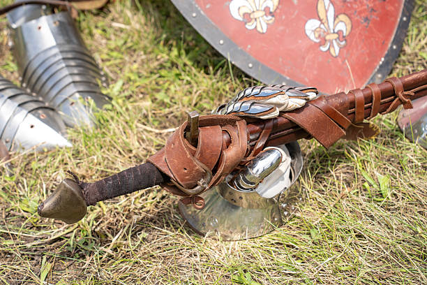 Close-up of a medieval sword and decorative shield resting on green grass, surrounded by steel gauntlets and armor pieces. The worn red and black shield with fleur de lis symbols and the leather-bound sword evoke historical reenactment and chivalric tradition.