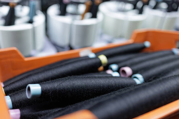 Closeup of black yarn cones in storage crate near automated textile machinery, illustrating industrial weaving preparation for fabric and apparel production marketing use.