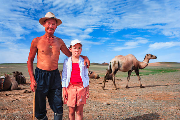 A shirtless older man wearing a wide-brimmed hat stands with his arm around a young girl on a dry, open plain. Several camels rest and one walks in the background beneath a bright blue sky dotted with clouds. The man holds a wooden staff, and the scene conveys a quiet moment of pastoral life in a sunlit desert landscape.