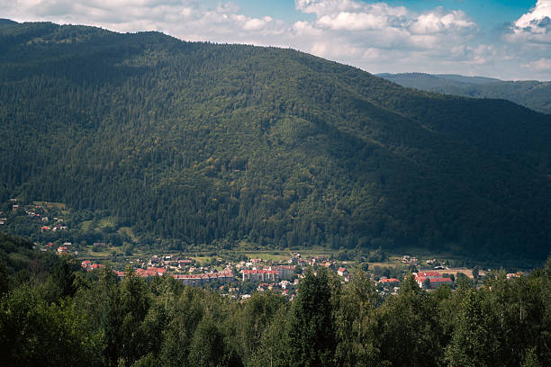 Panoramic view of green forested mountains with a small village in the valley under a blue sky with clouds. Scenic summer landscape, natural background ideal for travel, tourism, environment, and outdoor concepts.