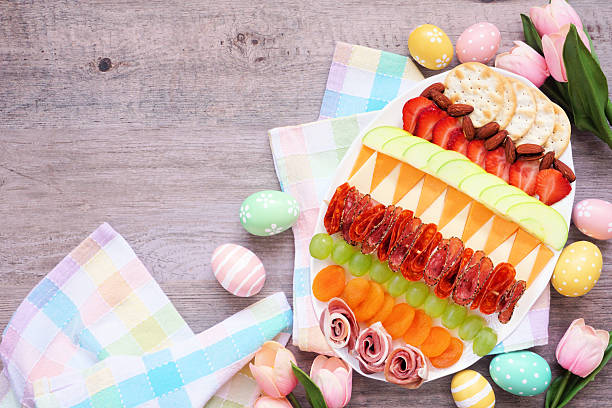 Fun Easter theme charcuterie board in a the shape of an egg. Top view table scene against a wood background. Variety of cheeses, meats and fruit.