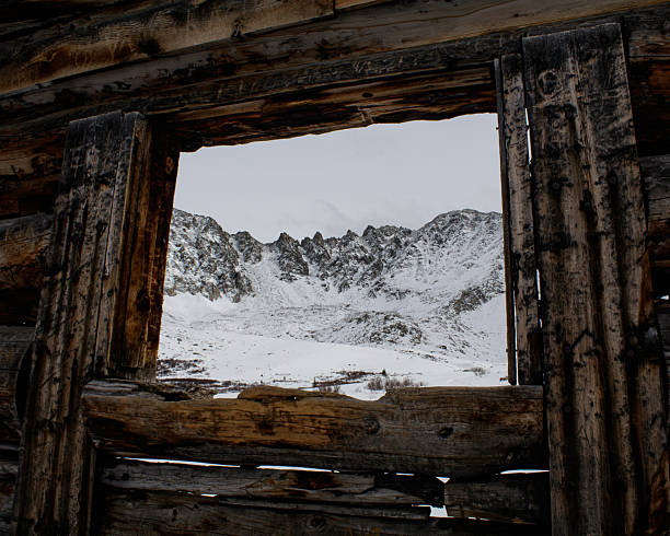 Snow covered alpine peaks viewed through the weathered wooden frame of an old cabin or mining structure. The rustic timber foreground contrasts with the cold winter landscape beyond, creating a natural frame and strong sense of depth. Ideal for themes of wilderness, solitude, exploration, history, and mountain adventure.