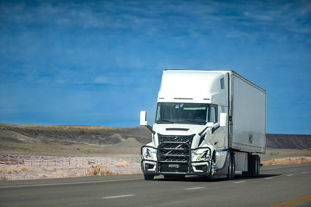 A white commercial semi-truck with a large trailer driving on an empty desert highway. The vehicle travels through an arid landscape with rocky terrain and clear blue sky, representing long-distance transportation and logistics industry.