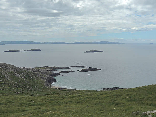 Coastal landscape with grassy hills, rocky shoreline, calm sea, and distant islands under a cloudy sky, creating a peaceful natural seascape with ample copy space.