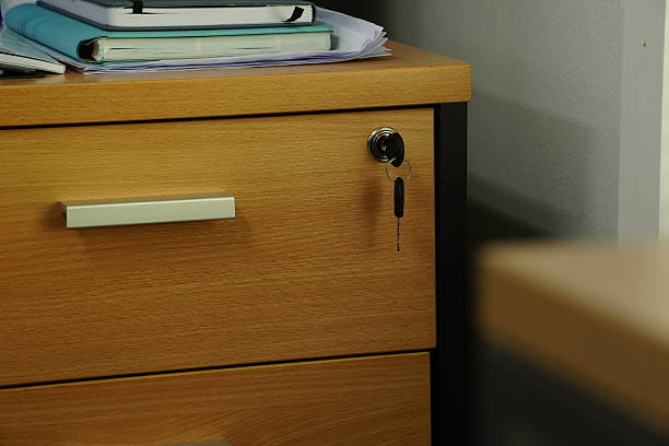 Soft, diffused natural light illuminates a wooden office desk, highlighting the simple details of a locked drawer and a stack of notebooks.