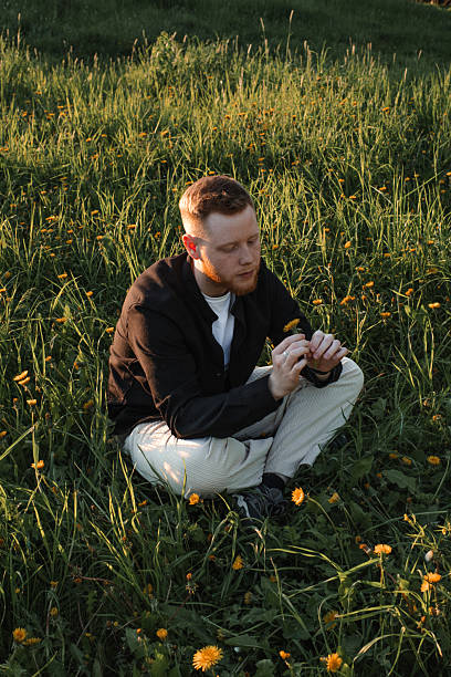 A young red-haired man with a beard sits on a blooming meadow at sunset.