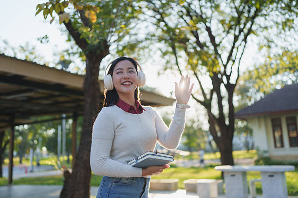 Young asian woman wearing headphones and holding books, waving hello with a smile outdoors