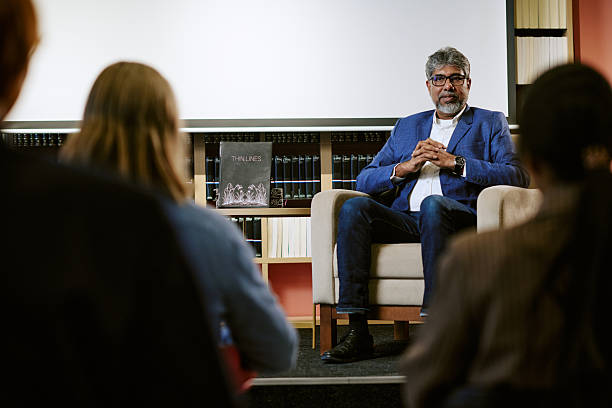 Middle aged man sitting on chair presenting to group of people in front of large screen, holding hands together, audience listening attentively during book presentation