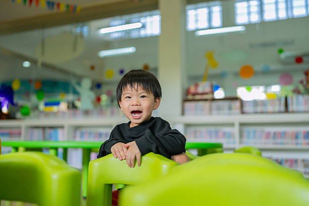 A joyful Asian toddler boy, around 2–3 years old, is smiling brightly while standing behind colorful green chairs in a vibrant children’s library or classroom. He is wearing a black long-sleeve shirt and appears excited and playful. The background features bookshelves filled with children’s books and bright decorative elements, creating a lively and educational atmosphere. The natural light and colorful surroundings highlight early childhood happiness, social development, and a positive learning environment that supports cognitive and emotional growth.