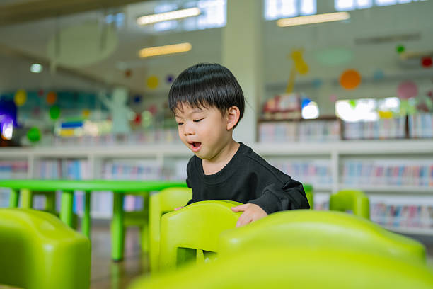 A joyful Asian toddler boy, around 2–3 years old, is smiling brightly while standing behind colorful green chairs in a vibrant children’s library or classroom. He is wearing a black long-sleeve shirt and appears excited and playful. The background features bookshelves filled with children’s books and bright decorative elements, creating a lively and educational atmosphere. The natural light and colorful surroundings highlight early childhood happiness, social development, and a positive learning environment that supports cognitive and emotional growth.