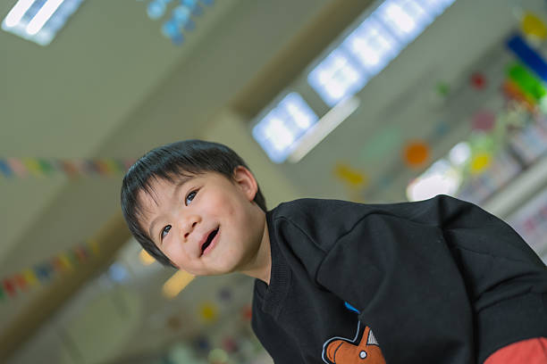 A joyful Asian toddler boy, around 2–3 years old, is smiling brightly while standing behind colorful green chairs in a vibrant children’s library or classroom. He is wearing a black long-sleeve shirt and appears excited and playful. The background features bookshelves filled with children’s books and bright decorative elements, creating a lively and educational atmosphere. The natural light and colorful surroundings highlight early childhood happiness, social development, and a positive learning environment that supports cognitive and emotional growth.