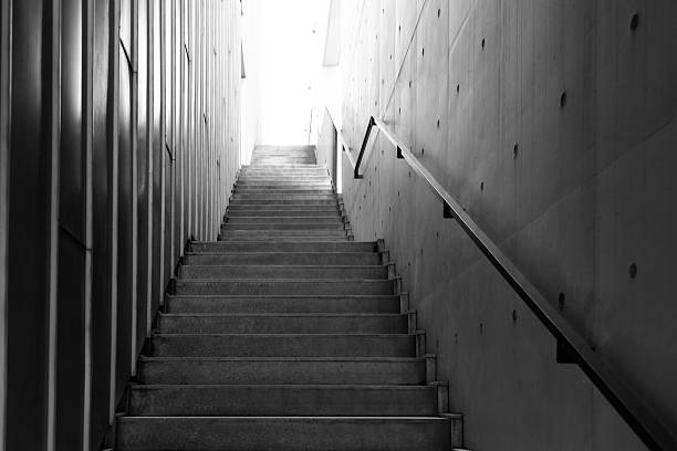 The image depicts a dimly lit stairway descending into an underground passage. The stairs are made of concrete or stone, with metal railings on one or both sides. The lighting is minimal, creating deep shadows and a sense of mystery or isolation. The walls appear rugged or industrial, possibly part of a basement, tunnel, or utility corridor. The overall atmosphere is dark and moody, suggesting an urban or hidden environment, often associated with maintenance areas, subways, or old architectural structures.