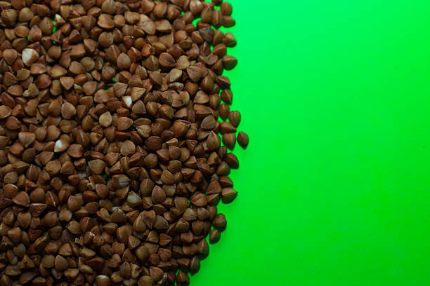 Dry buckwheat groats are scattered in an even layer on a vibrant lime green background, creating a distinct color contrast. This clean and minimalist image of a healthy cereal is perfect for visualizing healthy nutrition, diet recipes,