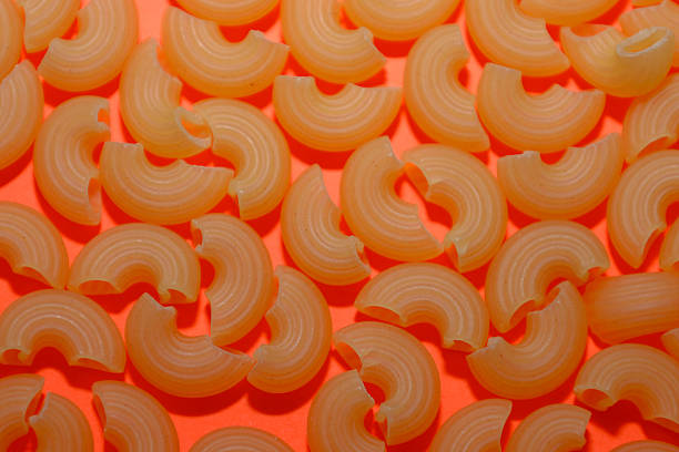 Dry elbow pasta tightly scattered on a vibrant orange background, creating a warm and appetizing color palette. This textured image is perfect for packaging design, culinary website decoration, or visualizing the theme of quick meal preparation.