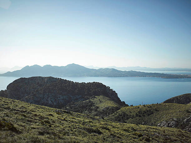 grassy hill looking out at the distant mountains and sea under a clear sky. The sun shines brightly over the landscape.