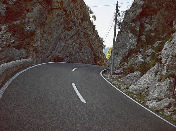 The scene shows a twisting road surrounded by tall rock formations. A single power line runs alongside the road. The sun shines down on the landscape, highlighting the rocky cliffs nearby.