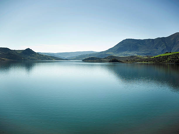 A large body of water reflects the blue sky with mountains surrounding the area. Green hills can be seen in the distance. It is daytime and the scene is clear.