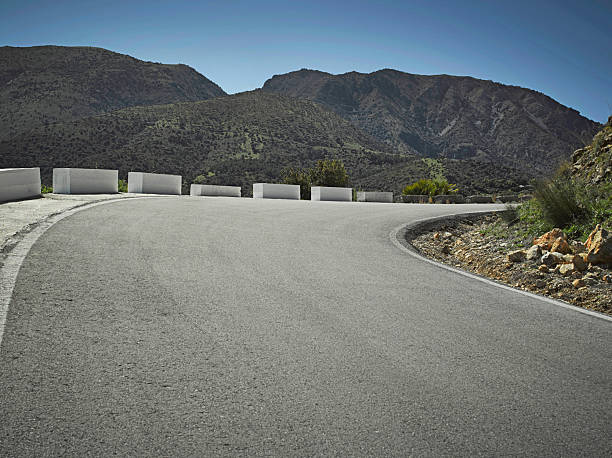 A road curves around mountains, marked by barriers on each side. The sun shines in a clear blue sky. The landscape shows hills and greenery in the background.