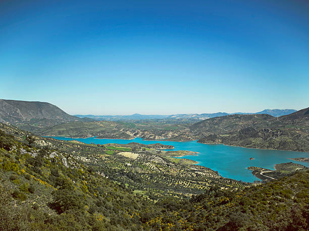 A wide view shows a lake surrounded by hills and mountains. The clear blue sky above the landscape highlights the colors of the water and greenery below.