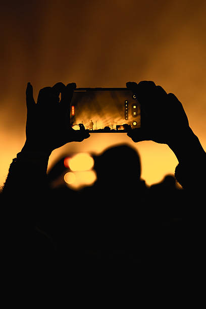 A person uses both hands to stabilize a mobile phone while filming a live stage show. The vibrant yellow and orange background heavily silhouettes the subject.