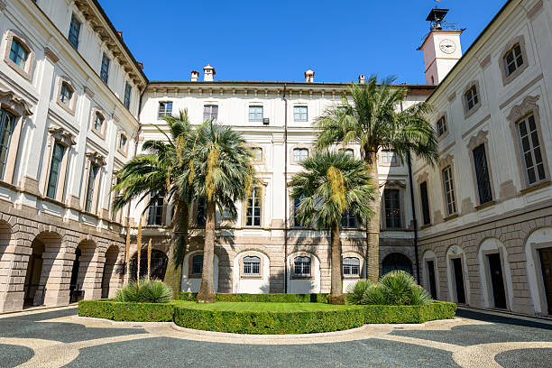 Isola Bella, Italy - September 30, 2020: The inner courtyard of the Palais Borromee features several palm trees, manicured shrubs, and a decorative patterned stone pavement surrounded by neoclassical palace facades under a clear blue sky.