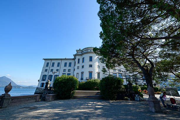 Isola Bella, Italy - September 30, 2020: The exterior of the Palais Borromee is shown with a waterfront staircase, large pine tree, gravel promenade, and several people sitting or standing under a clear blue sky.
