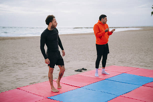 Two athletic male friends preparing for a brazilian jiu jitsu session on red and blue mats at a sandy beach
