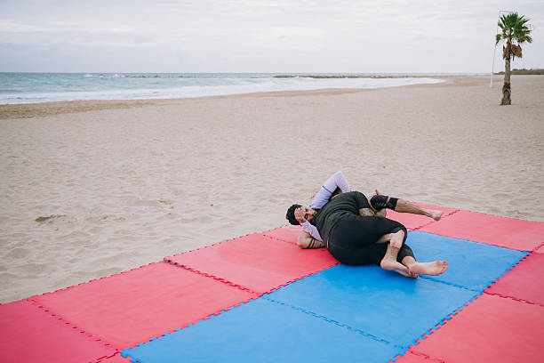 Male athlete practicing jiu jitsu grappling techniques on red and blue mats at a sandy beach with the ocean in the background