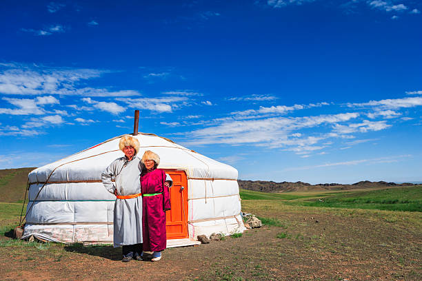 Two people dressed in traditional Mongolian robes and fur hats stand arm-in-arm in front of a white yurt with an orange door. They are positioned on a grassy steppe under a deep blue sky streaked with thin clouds, with low rolling hills visible in the distance. The scene is brightly lit by sunlight, showing vivid colors and a wide, open landscape.