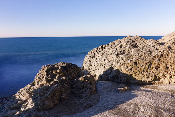 Bright and detailed shot of porous volcanic-style rocks on the shore with a smooth, long exposure sea in the background.