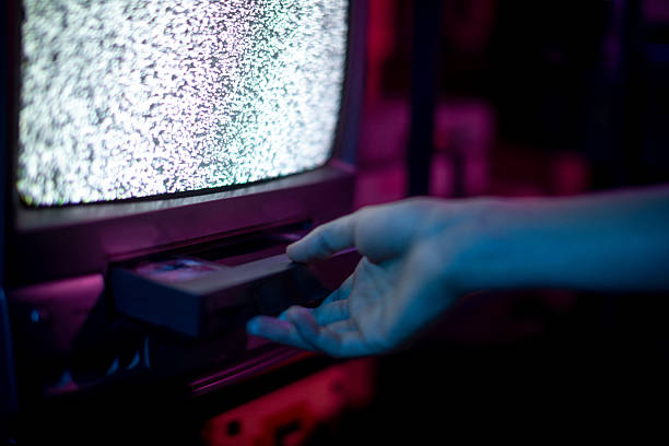 Cinematic close-up of a hand inserting a VHS cassette into a vintage CRT television displaying static noise. The neon color contrast enhances the retro atmosphere, evoking nostalgia, analog media culture, and pre-digital entertainment. Ideal for concepts related to retro technology, media history, nostalgia marketing, content consumption, and cultural storytelling around analog-era entertainment.