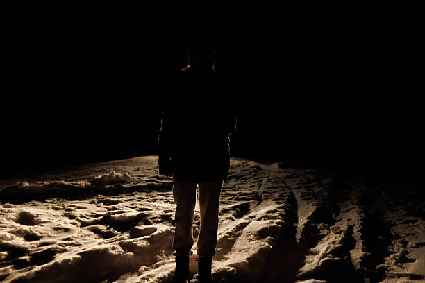 A person stands alone on a snowy surface as light shines on the ground. The surrounding area is dark with only the snow visible in the light. It is night in a winter setting.
