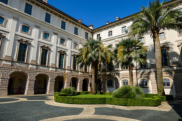 Isola Bella, Italy - September 30, 2020: The inner courtyard of the Palais Borromee features several palm trees, manicured shrubs, and a decorative patterned stone pavement surrounded by neoclassical palace facades under a clear blue sky.
