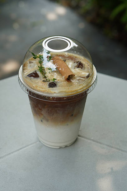 top view cool brown and white espresso coffee topped with layer and sprig of herbs and cinnamon stick in a clear plastic cup on white table, blur cement floor and natural background, drink,  copy space