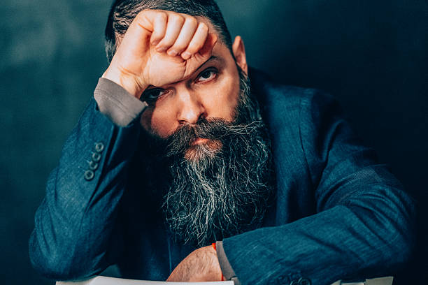Serious bearded man in a dark textured suit leaning his head on his fist against a dark studio background, expressing deep contemplation, concentration, or pondering business decisions