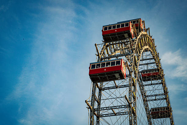 This photograph captures the upper half of a large Ferris wheel silhouetted against a soft, pale sky, likely at dawn or dusk. The wheel's white metal structure, consisting of the outer rim, supporting spokes, and a tall central mast with a lightning rod, dominates the frame. Spaced evenly along the rim are enclosed passenger cabins in varying shades of yellow, blue, and red. The simple composition and muted lighting create a serene and slightly nostalgic atmosphere.