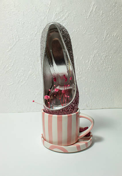Pink glitter stiletto high heel paired with striped coffee cup, sugar cubes and delicate flowers on white background. Feminine fashion and lifestyle concept.