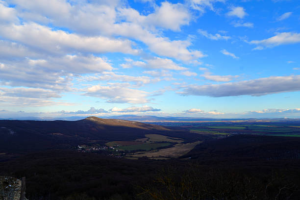 A group of hills lies in the distance. Green fields and small houses spread out below. The sky is bright with clouds in various shapes. It is daytime with clear visibility.
