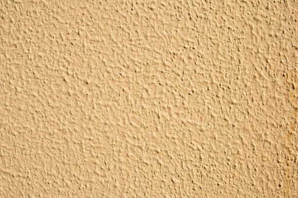 A detailed view of a rough, stucco-like wall in a warm beige hue, showcasing its uneven texture and natural light play