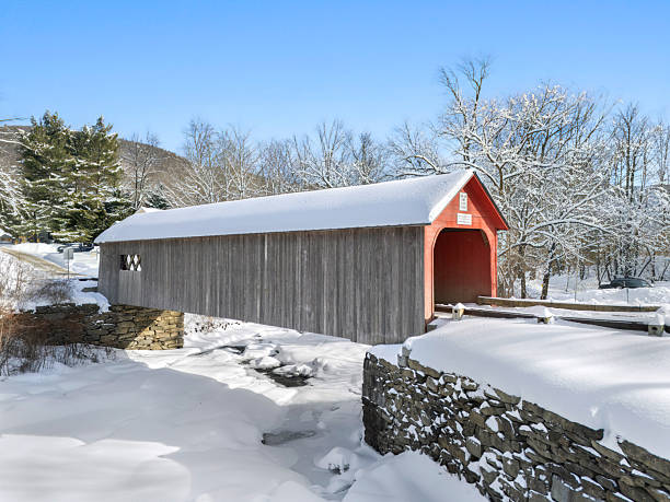 Wooden covered bridge with red entrance surrounded by snow under blue sky in rural New England, USA.