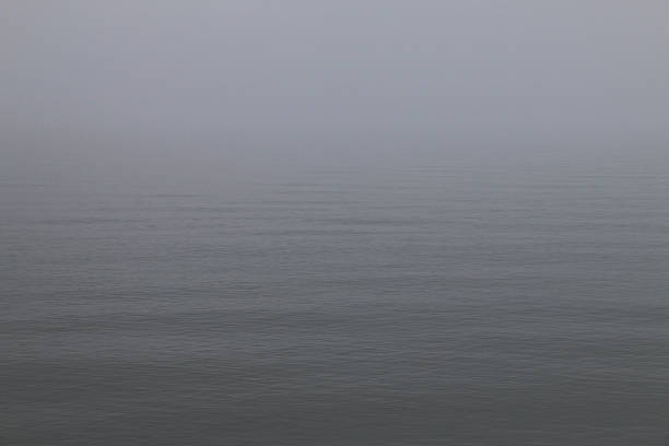 Expansive calm water under dense fog. Horizon barely visible, creating a minimalistic, tranquil scene with soft, muted tones and gentle ripples. Peaceful, introspective atmosphere.